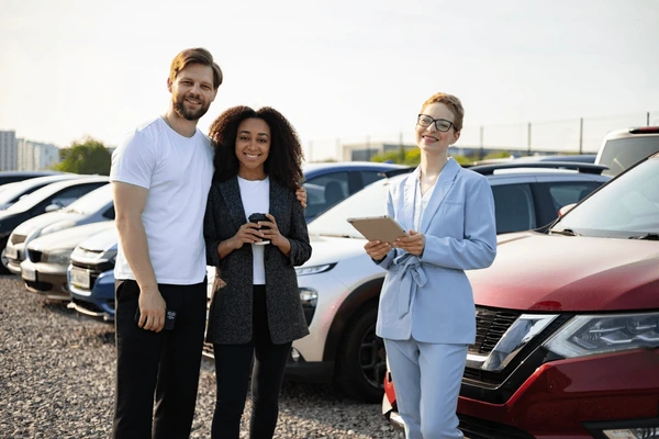 A friendly insurance agent handing a digital tablet to a smiling couple sitting in their new car
